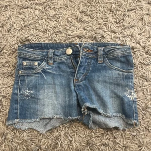 Blå jeansshorts från Detroit - Snygga blå jeansshorts från Detroit med fransig kant och slitna detaljer. Klassisk femficksmodell med knapp och dragkedja framtill samt coola bakfickor med lock och knapp. Perfekt för chill dagar och sommarhäng. Detta är barn shorts så därför väldigt små!