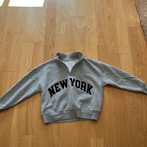 Grå half zip sweatshirt från Zara - Grå sweatshirt från Zara med svart 'NEW YORK'-tryck på bröstet. Tröjan har en cool half zip-dragkedja vid halsen och är långärmad med ribbade muddar. Mjuk insida i fleece och avslappnad passform, perfekt för chill dagar.