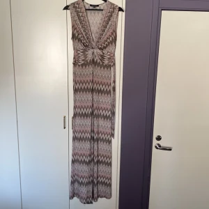 Mönstrad långklänning Ilse Jacobsen - Säljer en snygg långklänning från Ilse Jacobsen i storlek M. Klänningen har ett zigzag-mönster i beige, brun och rosa toner, omlott-v-ringad front och knyt i midjan. Ärmlös design och mjukt, stretchigt material som sitter bekvämt och ger en cool boho-vibe.