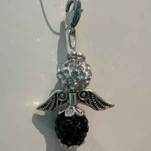 Glittrigt ängelhalsband med pärlor - Unikt halsband med ängelberlock i silverfärgad metall. Berlocken har en glittrig vit pärla upptill och en svart pärla nedtill, samt detaljerade vingar. Perfekt för dig som gillar statement-smycken med lite extra bling.