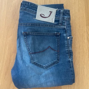 Jacob Cohen Jeans - Snygga blå jeans från Jacob Cohen med klassisk femficksdesign. Tillverkade i mjukt denimtyg med röda detaljer på bakfickorna. Det är en liten defekt på jeansen men inget mera. Priset är inte skrivet i sten!!!!!