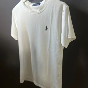 Vit t-shirt från Polo Ralph Lauren - Vit t-shirt från Polo Ralph Lauren med klassisk svart logobroderi på bröstet. Modellen är kortärmad och har rund hals. Tillverkad i mjuk bomull för skön känsla och clean look. Perfekt för dig som gillar stilrena och tidlösa plagg.