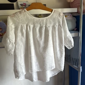 Vit blommig blus med broderi - Supersöt vit blus med broderade blommor och lätt transparent tyg upptill. Blusen har rund halsringning, korta ärmar och en luftig, flowy passform. Perfekt för en fräsch och romantisk look till sommaren eller våren!