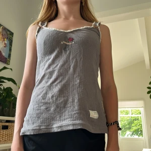 Grå ribbad linnetopp med rosett - Supersöt grå linnetopp med ribbad struktur och dekorativt tryck med röd ros framtill. Toppen har krämvit volangkant och en liten beige rosett på axelbandet. Materialet känns mjukt och luftigt, perfekt för varma dagar.