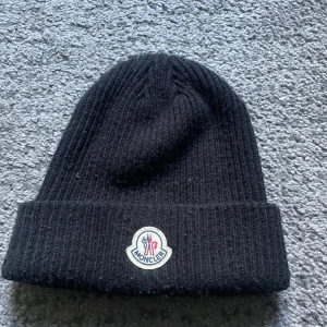 Svart ribbad mössa från Moncler - Svart ribbstickad mössa från Moncler med klassisk uppvikt kant och broderad logotyp framtill. Perfekt accessoar för kyliga dagar och ger en clean, stilren look.