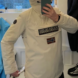 Napapijri Rainforest anorak - Snygg off white anorak från Napapijri med stor logga och norsk flagga på bröstet. Jackan har huva, dragkedja upptill och stor ficka framtill med lock. Insidan är fodrad med grått tyg för extra komfort och värme. Nypris ca 2500 kr. 