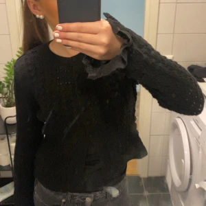 Svart stickad kofta med spetsdetaljer - Supersöt svart stickad kofta med genombrutet mönster och dekorativa knappar framtill. Ärmarna har volangkant i spets och samma spetsdetalj finns längs öppningen. Perfekt för dig som gillar romantiska och unika plagg.