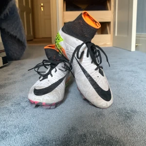 Nike Hypervenom fotbollsskor - Säljer ett par Nike Hypervenom fotbollsskor med högt skaft och snörning. Skorna är vita med svarta detaljer och har en färgstark sula i neonorange, rosa och gul med grafiskt mönster. Ovandelen är syntet och har en rund tå. Perfekt för dig som vill sticka ut på planen!