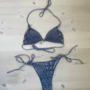 Blå mönstrad bikini med snörning - Snygg blå bikini med vitt mönster och knytband både upptill och nedtill. Bikinin har trekantsformad topp och fin snörning med en metall design på toppen. Perfekt för stranden och sommarens soliga dagar. Aldrig använd men utan prislapp.