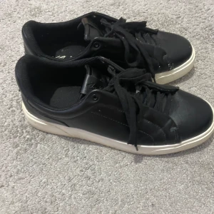 Svarta sneakers från pull&bear - Stilrena svarta sneakers från pull&bear med vit platt sula och klassisk snörning. Skorna har rund tå och är tillverkade i syntetmaterial med en enkel och clean look. Perfekta för dig som gillar minimalistisk stil och vill ha ett par bekväma vardagssneakers.