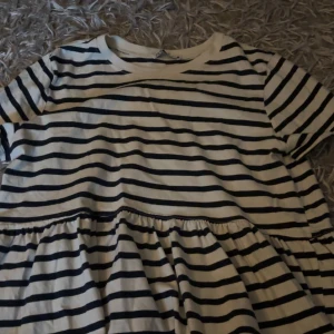 Randig T-shirt från Zara - Säljer en söt T-shirt från Zara i storlek S. T-shirten är vit med svarta horisontella ränder, har rund hals och korta ärmar. Den har en volangdetalj vid midjan som ger en avslappnad och trendig look. Perfekt för dig som gillar klassisk stil med en twist.