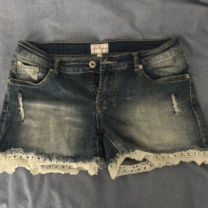 Blå jeansshorts med spets från Chica London - Snygga blå jeansshorts från Chica London med ljus tvätt och slitna detaljer. Shortsen har fem fickor och en cool spetskant i beige längs bensluten som ger en extra touch. Perfekt för dig som vill ha en chill och trendig sommarstil.