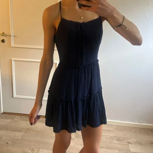 Marinblå kort klänning med knyt - Supersöt marinblå klänning med smala axelband och knyt framtill. Klänningen har volanger i kjolen, dekorativa knappar och en diskret dragkedja i sidan. Materialet är lätt och har små broderade prickar för extra detalj. Använd endast vid enstaka tillfälle då den var lite för kort för min smak. Jag är 174 för referens. Storlek 38 från märket Superdry. Nypris ca 800. 