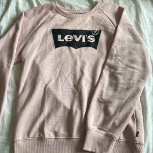 Ljusrosa Levi's sweatshirt med logga - Ljusrosa sweatshirt från Levi's med klassisk svartvit logga framtill. Tröjan har rund hals, långa ärmar och ribbade muddar. Perfekt för en chill och avslappnad stil. Tillverkad i mjuk bomull som känns skön mot huden. Finns små fläckar på den nedre ribbade delen och två små fläckar fram på bröstkorgen.