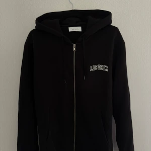Svart hoodie från Les Deux med dragkedja - Svart hoodie från Les Deux med vit broderad logga på bröstet. Tröjan har huva med snörning, praktisk dragkedja framtill och två fickor. Perfekt för chill dagar och enkel att matcha med jeans eller joggers.