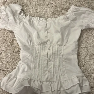 Vit korsettblus med volang  - Supersöt vit korsettblus från marknad med puffiga offshoulder-ärmar, knappar framtill och volangkant nertill. Snygga sömmar ger en figurnära passform och romantisk vibe. Perfekt för dig som vill sticka ut med en feminin och trendig topp. Lite stor på mig som brukar ha xs