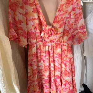 Blommig kortklänning med volang - Superfin kortklänning med blommigt mönster i rosa, orange och vitt. Klänningen har v-ringning med knyt framtill, volangdetaljer på ärmar och nederkant samt en luftig passform. Perfekt för soliga dagar och ger en fräsch vibe.