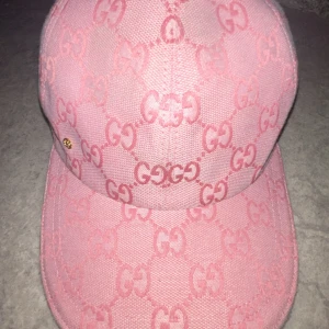 Rosa Gucci keps med GG-mönster - Snygg rosa keps från Gucci med ikoniskt GG-mönster över hela kepsen och en liten guldfärgad GG-detalj på sidan. Kepsen är tillverkad i bomull och har en klassisk böjd skärm. Perfekt för dig som vill sticka ut med en lyxig och trendig accessoar.
