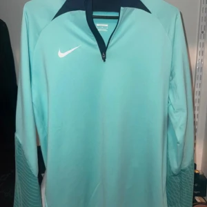 Nike Dri-Fit turkos träningströja - Nike Dri-Fit långärmad träningströja i turkos med mörkblå detaljer vid kragen och half zip. Slim fit-modell med coolt mönster på ärmarna. Perfekt för dig som vill ha en sportig och snygg look under träningen.