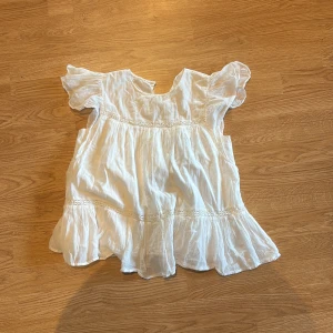 Vit blus med volang från Gina Tricot - Supersöt vit blus från Gina Tricot med volangdetaljer och korta ärmar. Blusen har en luftig och flowy passform med fina spetsinslag och romantisk känsla. Perfekt för varma dagar när du vill ha något lätt och stilrent.