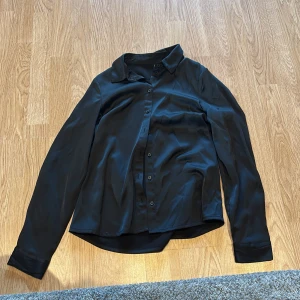 Svart skjorta från Vero Moda XS - Stilren svart skjorta från Vero Moda i storlek XS. Skjortan har klassisk krage, långa ärmar och knäppning framtill med svarta knappar. Materialet är mjukt och har en lätt glansig finish som ger en snygg look. Perfekt för dig som gillar minimalistisk stil.