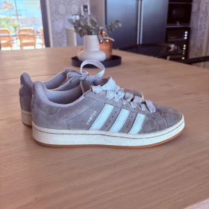Grå Adidas Campus sneakers i mocka - Snygga Adidas Campus sneakers i grå mocka med klassiska vita ränder och vit sula. Skorna har rund tå, platt sula och breda vita skosnören. Säljer då jag inte använder dessa längre.