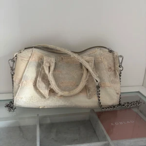Väska från Zara - Snygg beige handväska i jeansmaterial med slitna detaljer och silverfärgad kedja som axelrem. Väskan har två handtag och en stor dragkedja upptill. Perfekt för dig som gillar en cool och avslappnad stil med lite edge. Aldrig använd, och är i väldigt bra skick. 