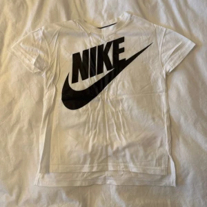 Vit Nike t-shirt med stor logga - Vit t-shirt från Nike med en stor svart logga tryckt på framsidan. Klassisk rund hals och korta ärmar. Tillverkad i mjuk bomull som är skön mot huden. Perfekt för dig som gillar sportig och enkel stil.