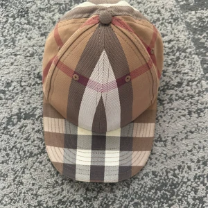 Burberry keps - Snygg keps från Burberry i klassiskt rutigt mönster med beige, bruna och röda toner. Kepsen har justerbart spänne bak och broderad Burberry-logga. Tillverkad i bomull och perfekt för dig som vill ha en stilren accessoar med lyxig känsla.