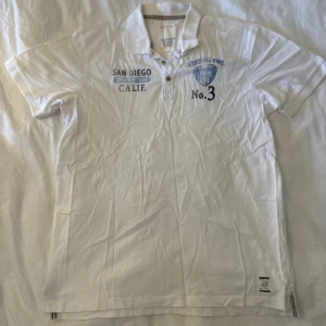 Vit polo t-shirt från Esprit - Vit polo t-shirt från Esprit med tryck på bröstet och coola festival-vibes. Har krage, två knappar och detaljer med blått och svart print. Materialet är mjuk bomull och t-shirten har korta ärmar. Snygg etikett nertill och slits på sidorna.