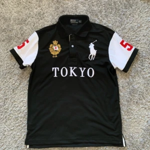 pikétröja Polo Ralph Lauren - Säljer en svart pikétröja från Polo Ralph Lauren med vita ärmar och stora röda siffror på ärmar och rygg. Framsidan har broderad logga, Tokyo-tryck och guldfärgad detalj. Klassisk krage och knappslå framtill.  rep