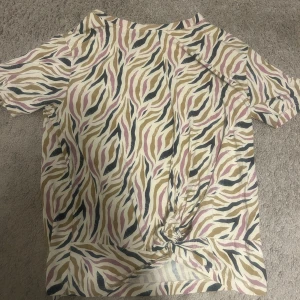 Zebramönstrad topp från The Nevi  - Cool zebramönstrad topp från The New i vitt, beige, rosa och svart. T-shirten har korta ärmar och en snygg knutdetalj framtill som ger en trendig look. Perfekt för dig som vill sticka ut med ett djurmönster och mjukt material.