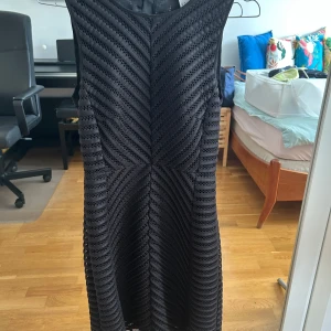 Svart ärmlös klänning från H&M - Snygg svart klänning från H&M i storlek 36. Klänningen är ärmlös och har ett coolt hålmönster med diagonala ränder. Den har en guldig dragkedja baktill och är figurnära med kort längd. Perfekt för dig som gillar stilrena detaljer och modern look.