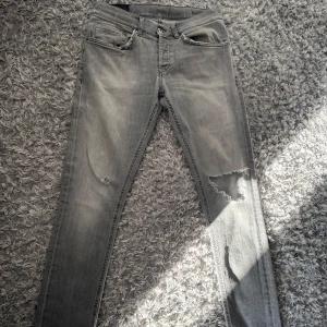 Grå skinny jeans med slitningar - Säljer ett par grå skinny jeans med snygga slitningar på ena benet och klassiska fem fickor. Jeansen har en smal passform och är tillverkade i mjukt denimtyg. Perfekta för dig som gillar en edgy och avslappnad stil.