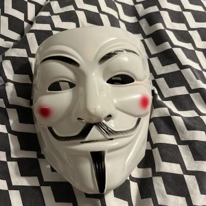 Vit Guy Fawkes mask med svart band - Säljer en vit Guy Fawkes mask med klassisk mustasch och leende, perfekt för maskerad eller temafest. Masken har ett svart elastiskt band som håller den på plats runt huvudet. Tillverkad i lätt syntetmaterial och passar de flesta huvudstorlekar.Ordinarie pris:100kr