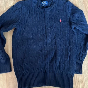 Mörkblå stickad tröja från Polo Ralph Lauren - Mörkblå kabelstickad tröja från Polo Ralph Lauren med rund halsringning och röd broderad logga på bröstet. Klassisk modell med ribbade muddar vid ärmslut och nederkant. Perfekt för en stilren look. Obs! Om ni ska köpa välj PostNord 