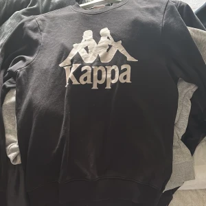 Svart sweatshirt från Kappa - Svart sweatshirt från Kappa med stort vitt logotryck på bröstet. Klassisk rund halsringning och långa ärmar. Perfekt för en avslappnad och sportig stil.