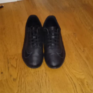 Svarta fotbollsskor från Nike - Säljer ett par helsvarta fotbollsskor från Nike med klassisk design och snörning. Skorna har rund tå och platt sula, perfekta för dig som vill ha en stilren look på planen.