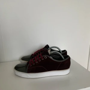 Lanvin skor - Säljer ett par vinröda sneakers från Lanvin med grå tå och detaljer samt vita sulor. Skorna har snörning och en klassisk rund tå. Perfekta för dig som vill ha en stilren och lyxig sneaker. Skicket är bra, tveka inte att höra av dig om du har frågor!
