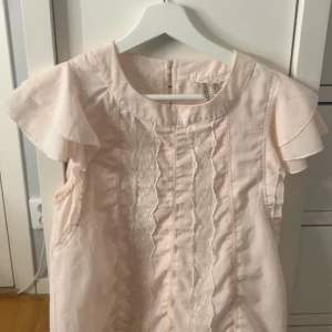 Ljusrosa blus med spetsdetaljer - Söt ljusrosa blus med volangärmar och dekorativa spetsdetaljer framtill. Blusen har rund halsringning och är tillverkad i ett lätt material, perfekt för varmare dagar. Storlek 38 men passar mig som är small/36 
