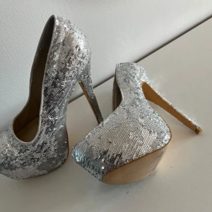 Silvriga glittriga pumps med paljetter - Säljer ett par höga pumps i silver med glittriga paljetter över hela skon. Skorna har en rejäl klack och plattform framtill, samt rund tå. Perfekta för att sticka ut på festen!