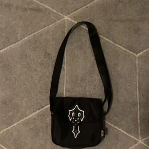 Svart axelväska från Trapstar - Säljer en svart axelväska från Trapstar med justerbart axelband och vit logga på locket. Väskan har ett unikt spänne framtill och är perfekt för att bära dina viktigaste saker. Snygg och stilren design med tydliga Trapstar-detaljer.