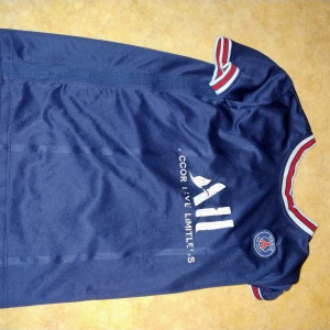 PSG Neymar fotbollströja - Mörkblå PSG fotbollströja med Neymar JR och nummer 10 på ryggen. Tröjan har vita tryck, klubbmärke på bröstet och röda, vita och blå detaljer vid ärmslut och krage. Perfekt för fans av Neymar och PSG.