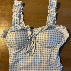 Blåvit rutig croppad topp med volang och knyt - Supersöt croppad topp i blåvitt rutigt mönster med volangdetaljer vid axelbanden och bysten. Toppen har en söt knytning framtill och breda axelband med volanger. Köpt på en mycket fin och dyr butik i Grekland! Perfekt för sommardagar!
