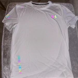  Under Armour Tshirt - Säljer en fräsch vit t-shirt från Under Armour med färgglada loggor och textdetaljer på bröstet, axeln och ärmen. Perfekt för träning eller sportiga outfits. Materialet känns lätt och andas bra. Perfekt för sommaren