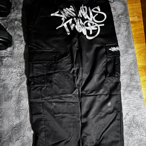 Svarta cargopants med vit graffiti-tryck - Svarta cargopants med stora vita graffiti-inspirerade tryck på framsidan och baksidan. Byxorna har flera fickor på sidorna och resår vid bensluten. Perfekta för en streetwear-look.