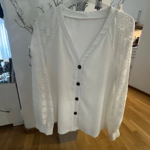 Vit kofta med spetsärmar och knappar - Säljer en vit kofta med v-ringning och svarta knappar framtill. Ärmarna är i genombruten spets med cirkelmönster som ger en snygg detalj. Perfekt att slänga över en topp för en fräsch look.
