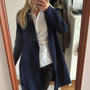 Mörkblå kappa med bälte från H&M - Säljer en stilren mörkblå kappa från H&M i ullblandning. Kappan har lång ärm, klassisk krage och knyts med bälte i midjan. Jag är egentligen en 38 så passar både 38/40!