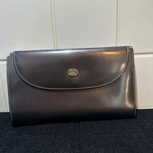 Mörkbrun plånbok i skinn från AGA - Säljer en stilren mörkbrun plånbok i skinn från AGA med guldfärgad logga på framsidan. Plånboken har flera fack för kort och mynt, samt en dragkedja inuti. Perfekt för dig som vill ha en klassisk och tidlös accessoar.