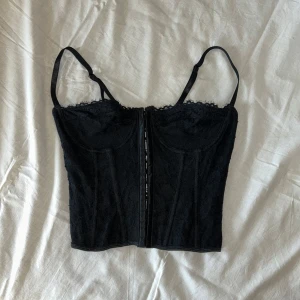 Svart spetskorsett med snörning - Säljer en svart korsett med spetsdetaljer och justerbara axelband. Framsidan har en dragkedja och baksidan har snygg snörning. Perfekt för att lyfta vilken outfit som helst. Helt oanvänd!💞